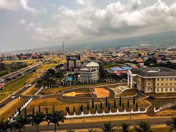 Vista aérea del ayuntamiento de Malabo, capital de Guinea Ecuatorial, con una plaza frente al edificio diseñada en forma de máscara tradicional, rodeada de cipreses y avenidas, con la ciudad y las colinas al fondo