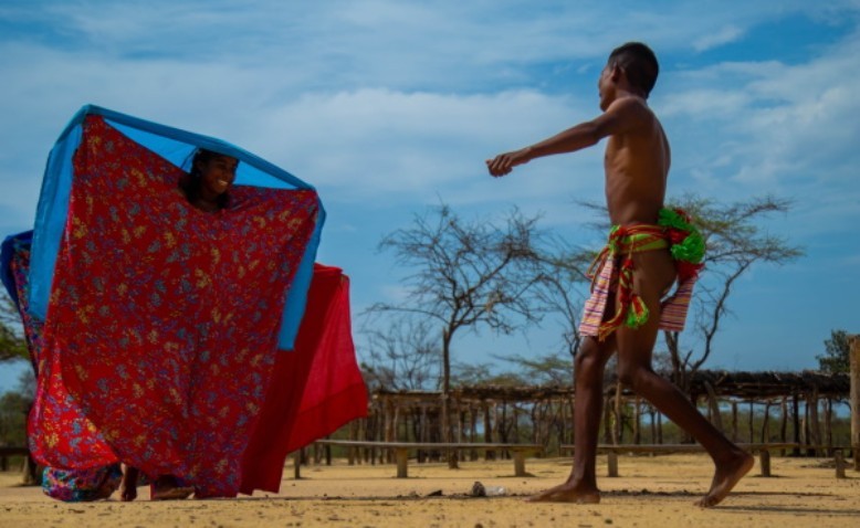 Mujer wayúu envuelta en una manta roja y azul durante la danza yonna, mientras un hombre con vestimenta tradicional baila frente a ella en un territorio semiárido